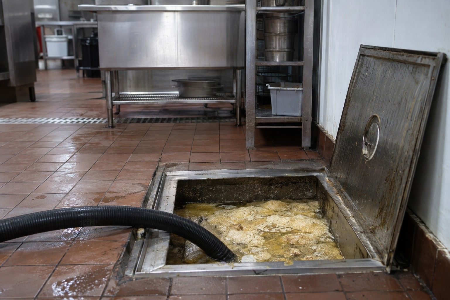 Bac à graisse ouvert dans le sol carrelé d'une cuisine de restaurant, couche de graisse figée et tuyau d'aspiration